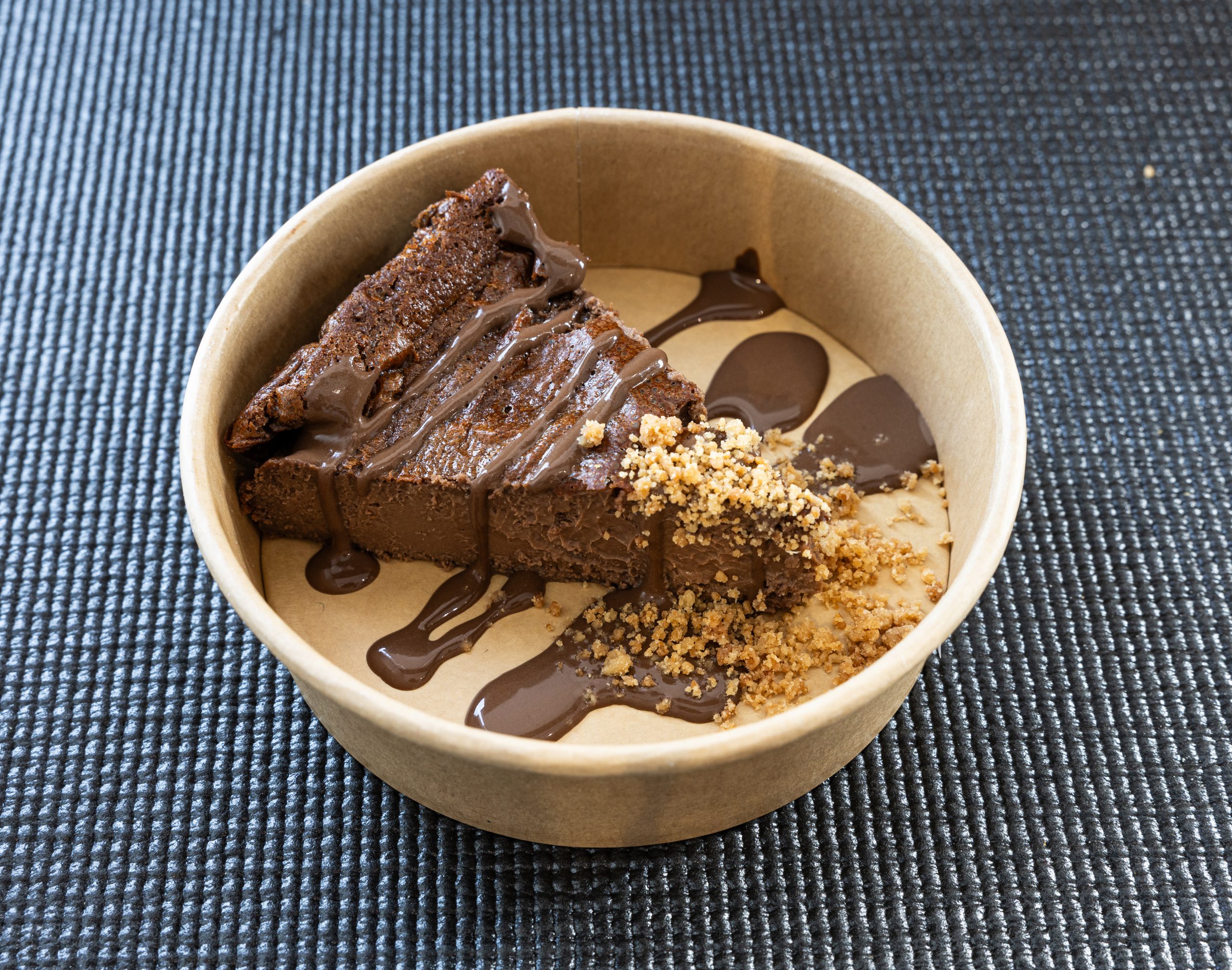 Porción de cheesecake de chocolate con salsa de chocolate y crumble servida en envase biodegradable